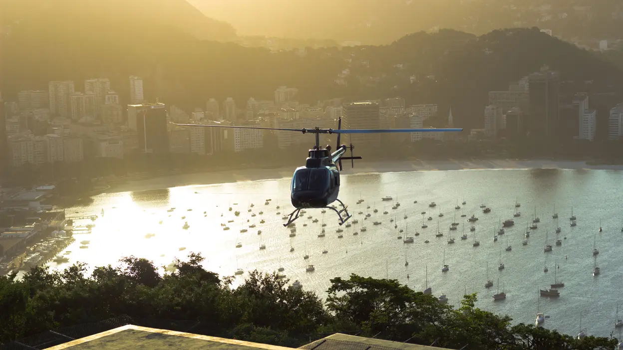 Helicopter flying over a coastal city with a bay full of anchored boats at sunset.