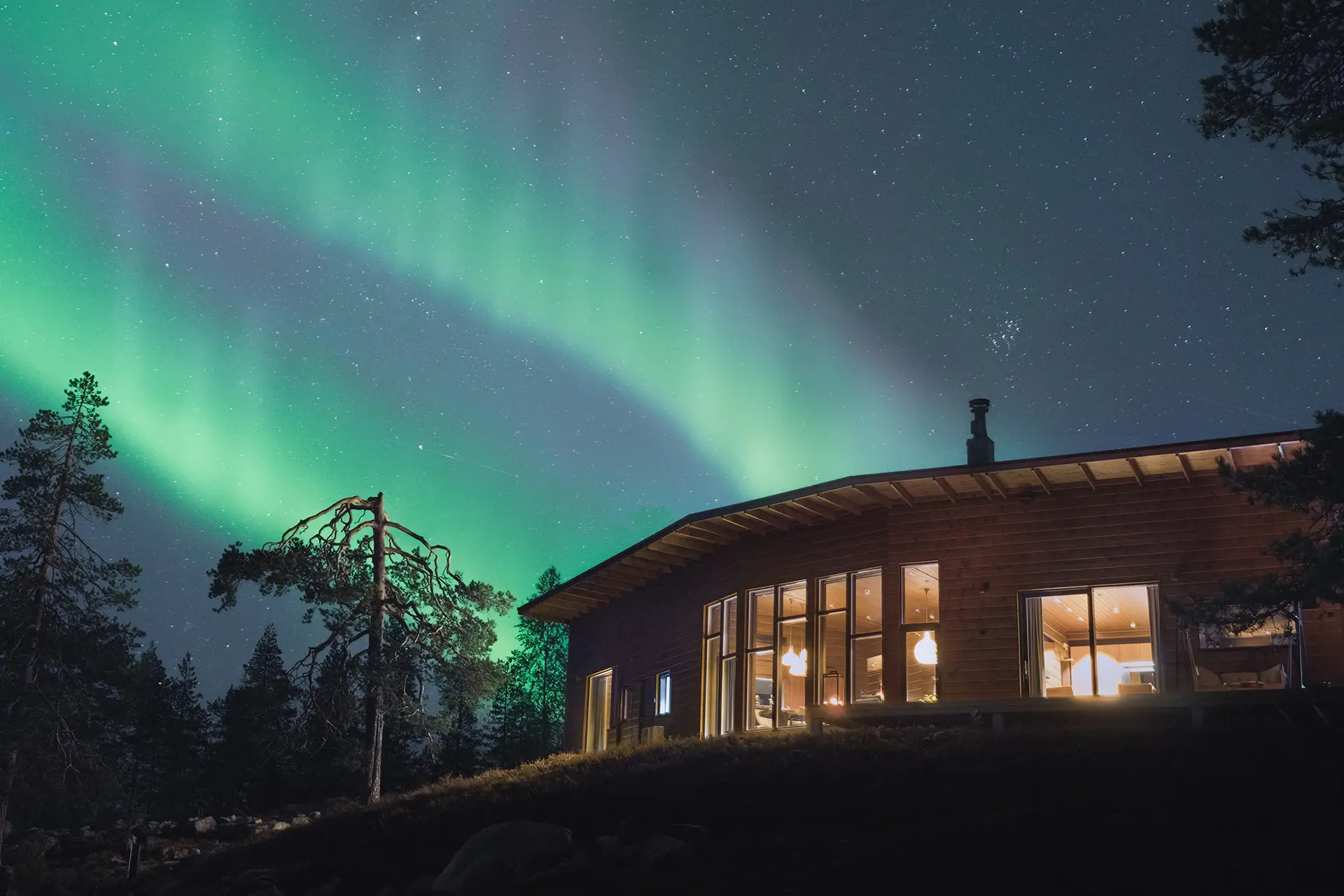 Octola Villa illuminated under the mesmerizing Northern Lights, surrounded by a starry sky and tranquil forest, offering a magical winter escape in the Arctic wilderness.