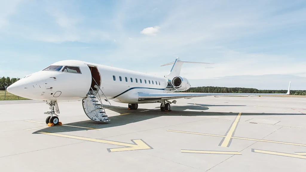 Featured aircraft from Avcon Jet: a stunning Global 5000 Vision private jet with a sleek white body and open staircase, parked on a sunny airport tarmac with a serene backdrop of trees and clear skies.